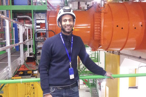 Dr Sudarshan Paramesvaran stands in front of a large room of equipment at CERN, wearing a hard hat.