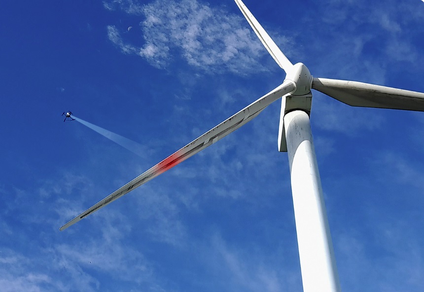 A drone flies near the blade of a wind turbine against a blue sky.