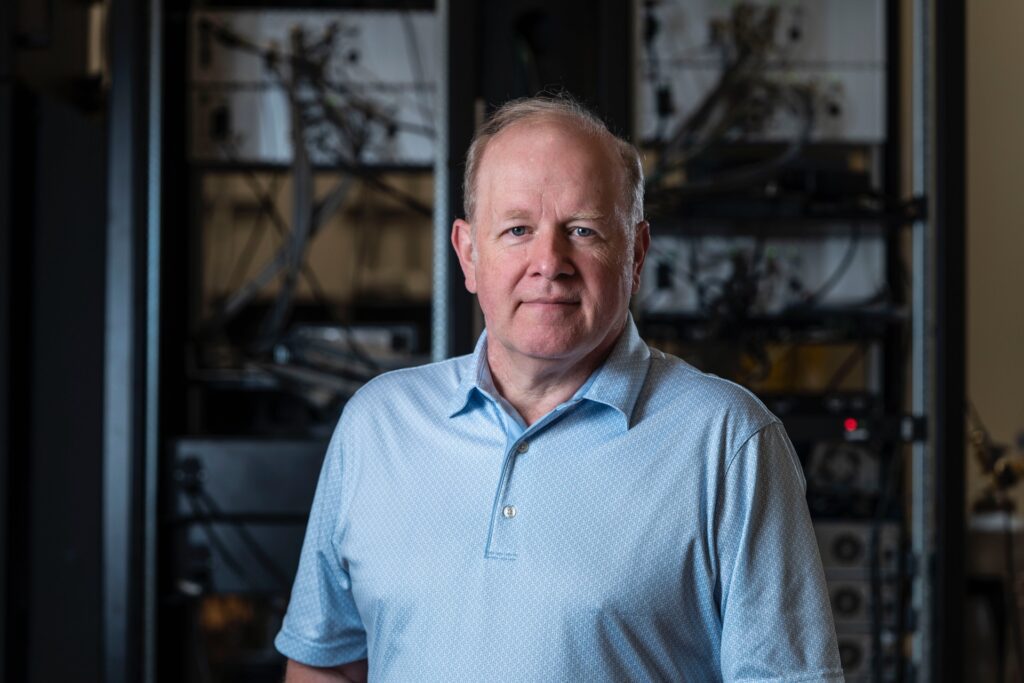 Peter Chapman of IonQ standing in front of an array of electronic equipment.