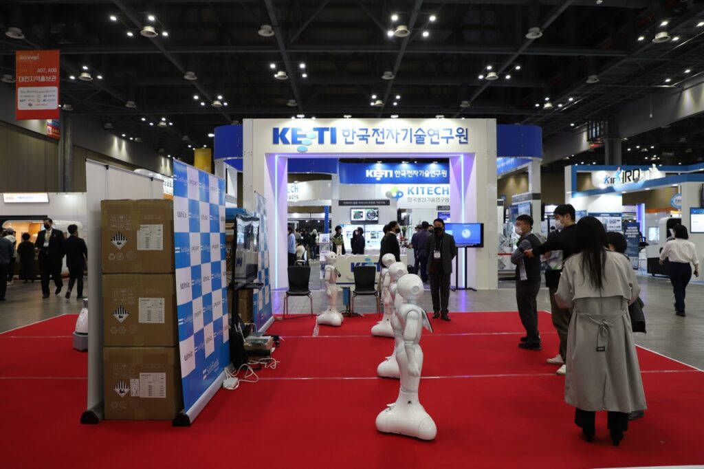 An image from a large robotics conferences, showing people walking past and looking at a line of small white robots.