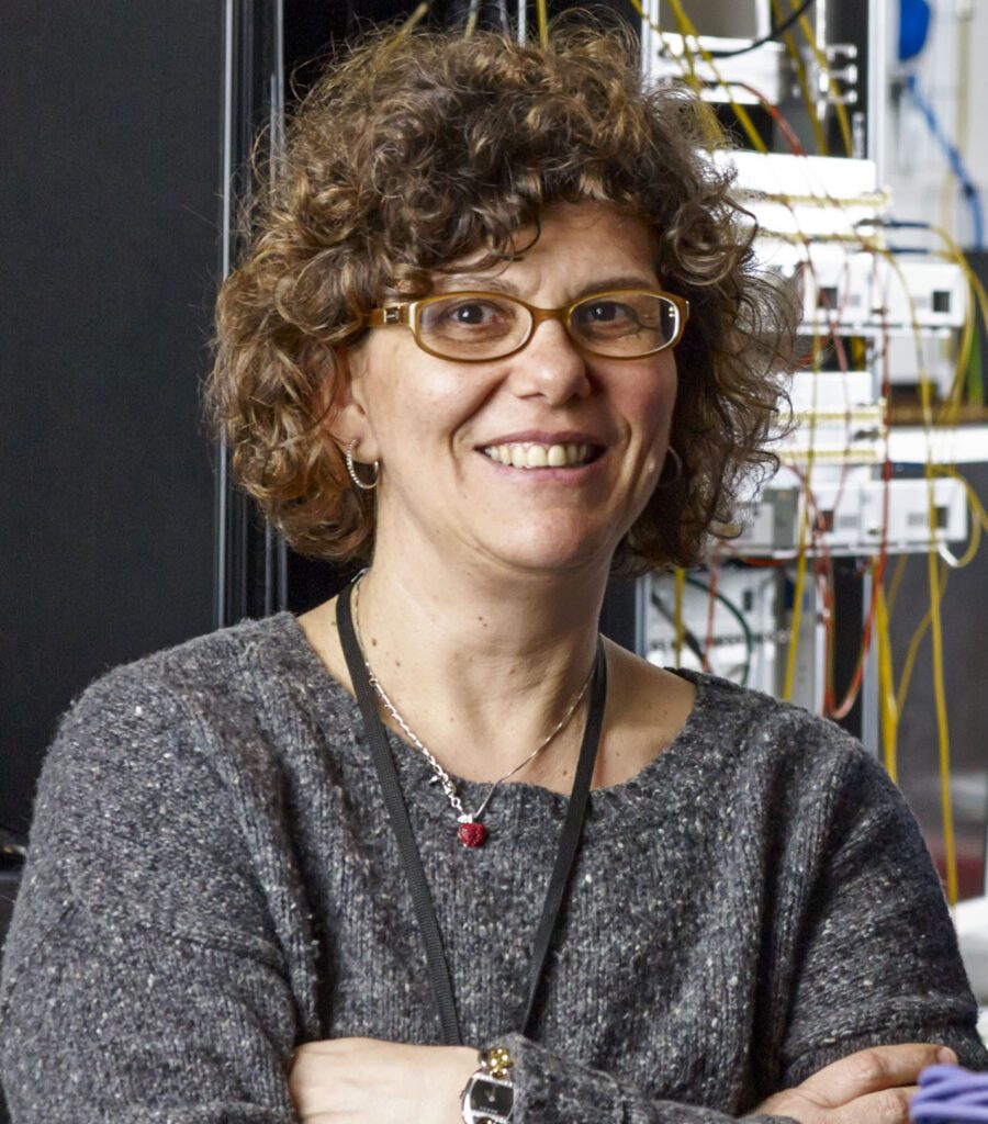 A headshot of Professor Dimitra Simeonidou, a woman with short curly hair and glasses, standing with electrical equipment in the background