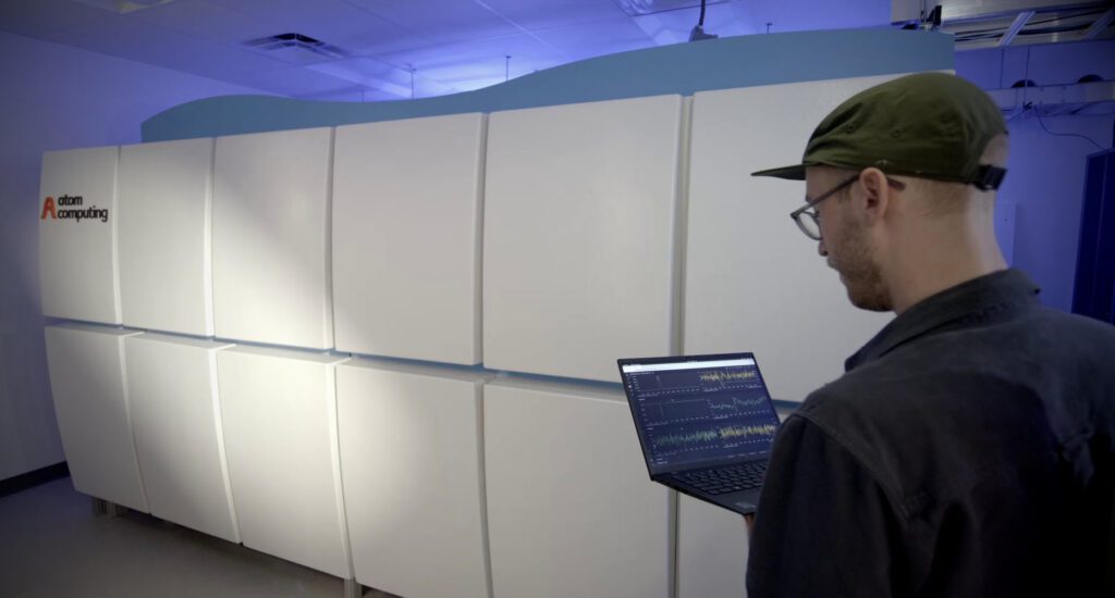 A man in a baseball cap holding a laptop in front of him stands in front of a bank of white facings, with Atom Computing written on the furthest one.