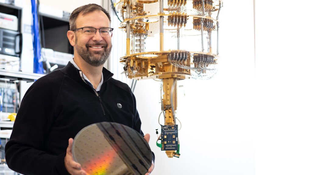 James Clarke, director of the Quantum Hardware research group within Intel’s Components Research Organisation, holding up a round disc while standing in front of electronic equipment.