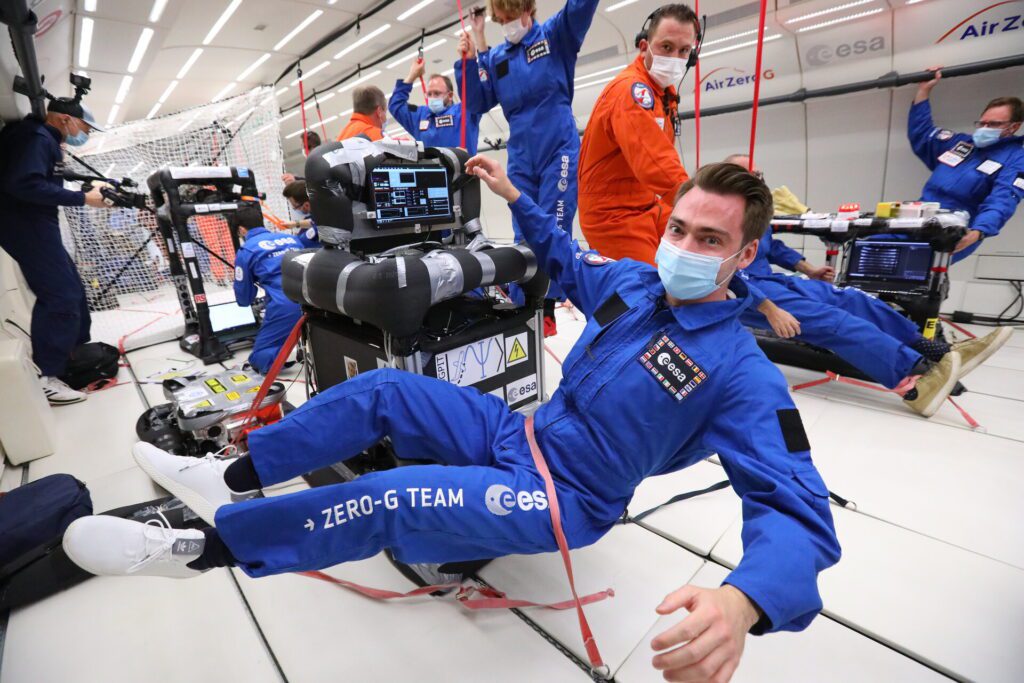 A group of scientists in blue and orange jumpsuits float in low gravity.