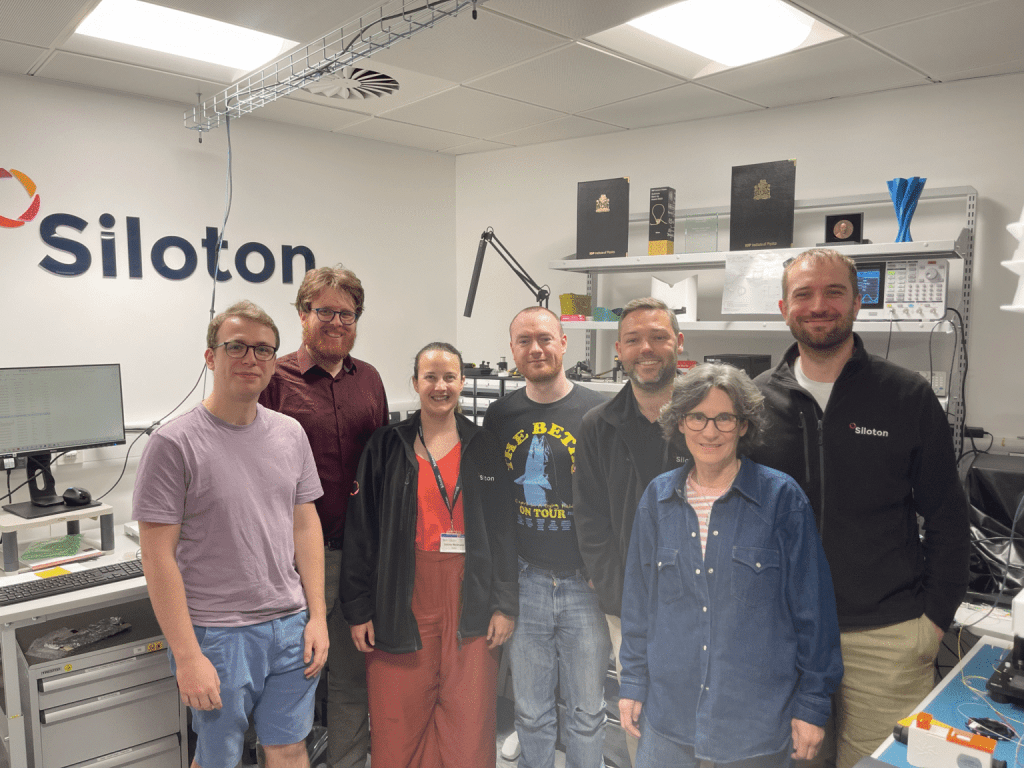 A group of people standing in a room filled with electrical equipment. A sign says Siloton on the wall behind them.