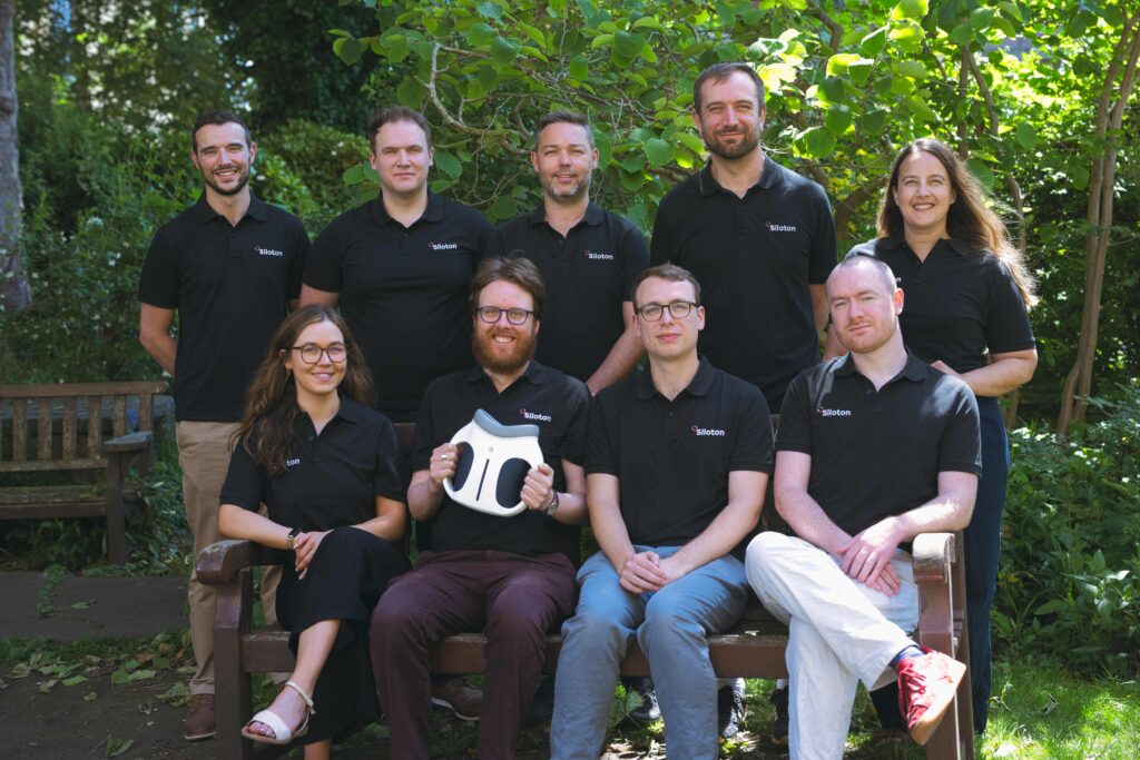 Nine men and women in matching black polo shirts with the Siloton logo on them. One is holding an optical device in front of his chest.